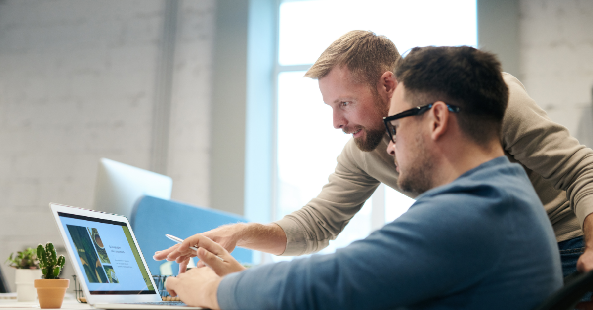 man using laptop at desk whilst another male colleague points to something on his screen 